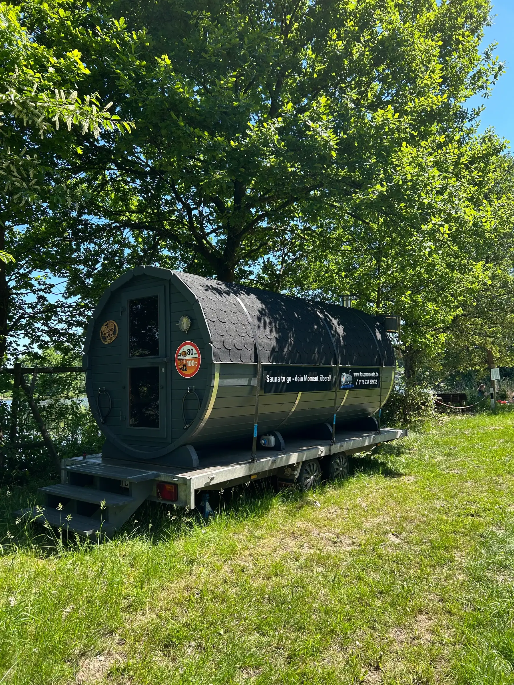 Schwarze XXL-Fasssauna auf dem Anhänger unter Bäumen